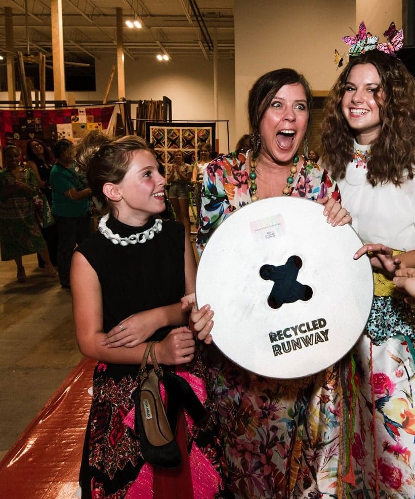 Three Recycled Runway participants shrieking with excitement as they hold up a giant Recycled Runway sign
