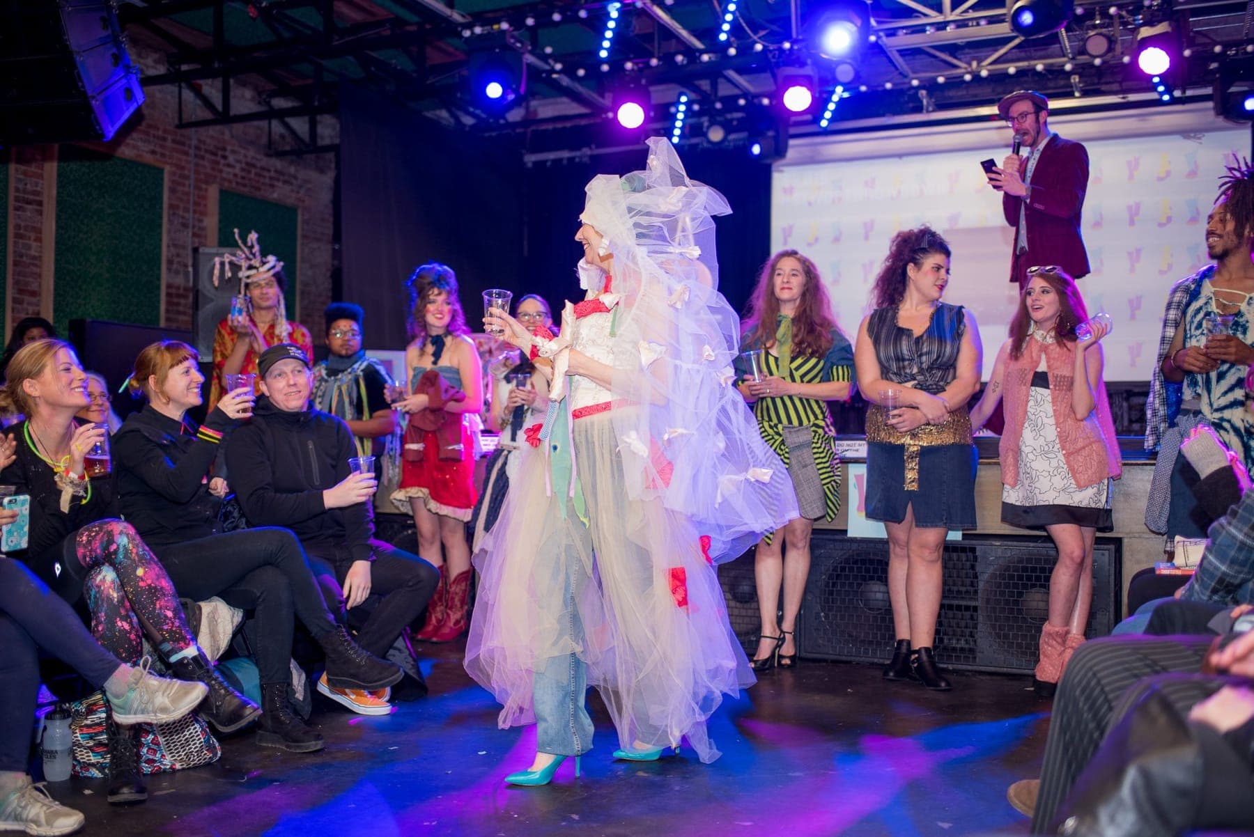 A model in a handmade white gown on stage while contestants and the audience watch